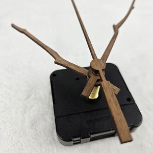 May include: A clock movement with wooden clock hands and a black plastic casing. The hands are a light brown color and have a unique branch-like design. The clock hands are attached to a brass-colored center.
