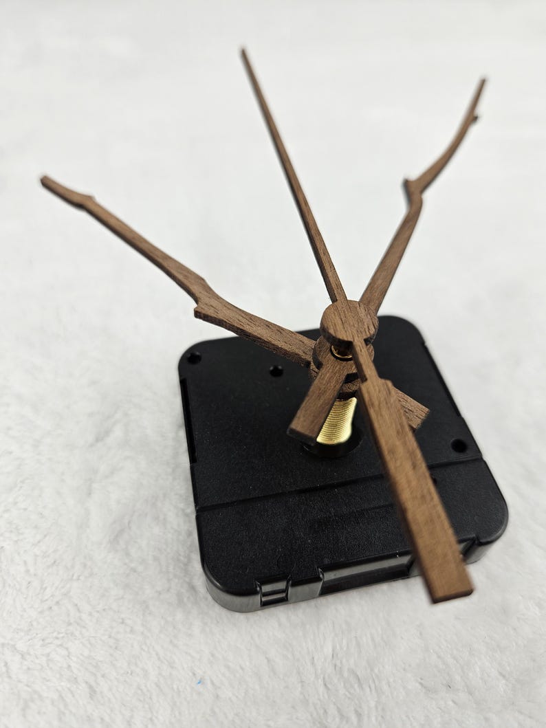 May include: A clock movement with wooden clock hands and a black plastic casing. The hands are a light brown color and have a unique branch-like design. The clock hands are attached to a brass-colored center.