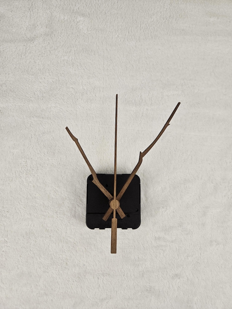 May include: A clock mechanism with wooden hands and a black square base. The hands are designed to resemble twigs and branches. The clock is set against a white, textured background.