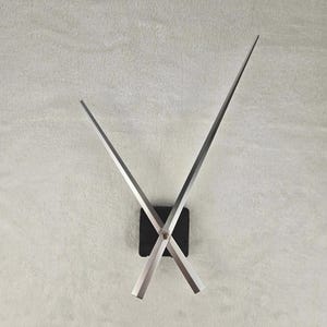 Könnte beinhalten: Eine moderne Wanduhr mit silberfarbenen Zeigern und einem schwarzen quadratischen Mittelteil. Die Zeiger sind lang und abgewinkelt und kreuzen sich in der Mitte. Die Uhr ist an einer hellgrauen, strukturierten Wand befestigt.