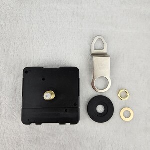 May include: Clock movement kit with a black square clock mechanism, a silver metal hanger, a black rubber washer, and two gold-colored nuts. The components are arranged on a light gray surface.