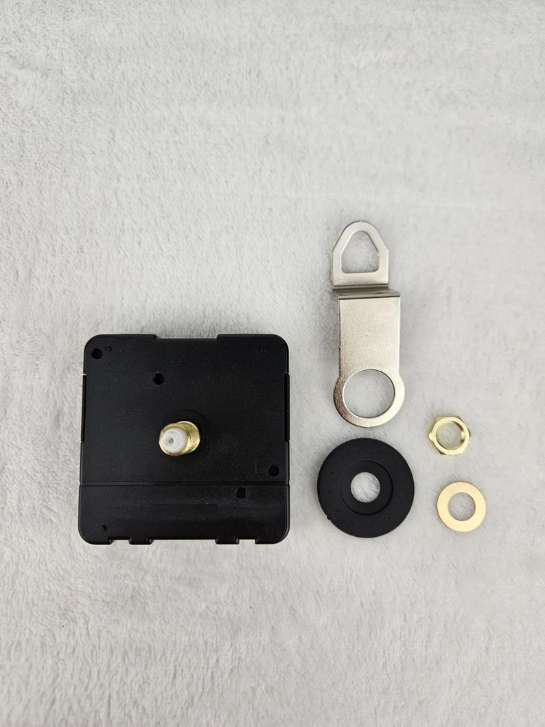 May include: Clock movement kit with a black square clock mechanism, a silver metal hanger, a black rubber washer, and two gold-colored nuts. The components are arranged on a light gray surface.