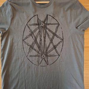 Op de afbeelding: Een grijs t-shirt met een zwart geometrisch ontwerp. Het ontwerp is een cirkelvormig patroon met elkaar kruisende lijnen, waardoor een stervorm ontstaat. Het shirt is gemaakt van een zacht materiaal en heeft korte mouwen.