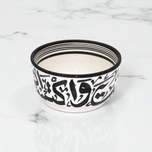 May include: A small, round ceramic bowl with a white interior and exterior. The rim is decorated with black stripes, and the exterior features black calligraphy. The bowl is likely used for serving food or as a decorative item.