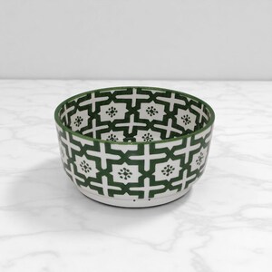 Peut inclure: Un bol en céramique blanc avec un motif géométrique vert. Le bol a une forme arrondie et une large ouverture. Le motif vert est composé de croix et de formes florales. Le bol convient pour servir des aliments ou comme objet décoratif.