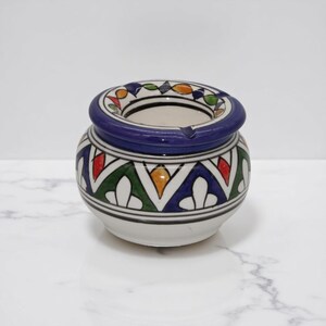 Peut inclure: Un cendrier en céramique avec une base blanche et un bord bleu. Le cendrier présente un motif géométrique en vert, rouge, jaune et bleu. Le design est symétrique et décoratif, avec une forme circulaire et une petite encoche.