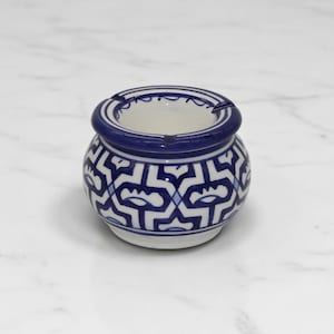 May include: A small, round ashtray with a blue and white geometric pattern. The ashtray has a wide, blue rim and a white interior. The exterior features a repeating design of stylized shapes in blue against a white background. The ashtray is made of ceramic.