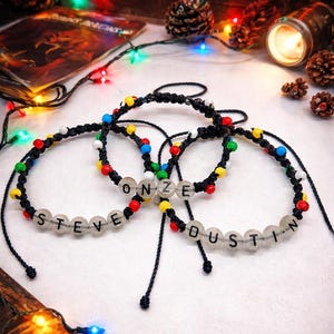 Könnte beinhalten: Drei schwarze Armbänder mit bunten Perlen und durchsichtigen Buchstabenperlen mit den Aufschriften "STEVE", "ONZE" und "DUSTIN". Die Armbänder werden mit festlichen Lichtern und Tannenzapfen präsentiert.