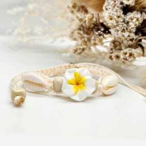 Könnte beinhalten: Ein Armband aus weißem Kordel mit einem weißen und gelben Blumenanhänger, weißen Muscheln und Holzperlen.
