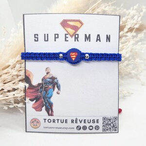 Op de afbeelding: Een blauwe macrame armband met zilveren kralen en een Superman logo bedel. De armband wordt getoond op een kaart met het woord "SUPERMAN" en een Superman illustratie. De kaart bevat ook de tekst "TORTUE RÊVEUSE".
