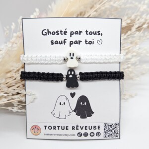 Könnte beinhalten: Zwei Makramee-Armbänder, eines weiß und eines schwarz, mit Geisteranhängern. Die Armbänder werden auf einer Karte mit dem Text "Ghosté par tous, sauf par toi" und einer Geisterillustration präsentiert. Der Markenname "TORTUE RÊVEUSE" ist ebenfalls sichtbar.