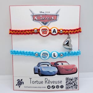 Op de afbeelding: Twee vriendschapsarmbanden met Cars-personages Bliksem McQueen en Mater. De rode armband heeft een letter A-bedel en de blauwe armband heeft een letter L-bedel. De armbanden bevinden zich op een witte kaart met het Cars-logo en de tekst "Tortue Rêveuse" en een website-adres.