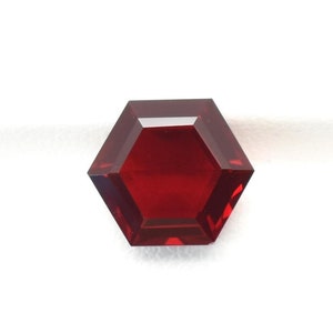 May include: A faceted, hexagonal gemstone with a deep red hue. The gem is set against a white background within a black display case. The cut of the stone allows light to reflect, creating a brilliant appearance. This gemstone is suitable for jewelry.