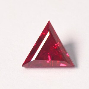 May include: A triangular-cut gemstone with a deep red color and faceted surfaces that reflect light. The stone features sharp, pointed corners and flat edges, displaying a translucent quality with internal light refractions throughout the body.