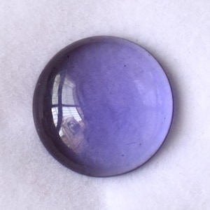 Peut inclure: Une pierre précieuse ronde et polie de couleur violette, présentée dans une boîte blanche. La pierre a une surface lisse et réfléchissante, légèrement bombée. La couleur est un violet profond et translucide.