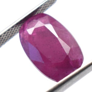 Op de afbeelding: Een gefacetteerde, ovaalvormige robijn edelsteen, vastgehouden door een zilveren pincet. De robijn heeft een diepe, verzadigde rode kleur met interne reflecties. De edelsteen staat tegen een heldere, witte achtergrond.