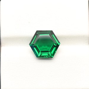Puede incluir: Una gema hexagonal de color verde esmeralda facetada. La gema se presenta sobre un fondo blanco dentro de un estuche negro cuadrado. Las facetas de la gema reflejan la luz, mostrando su claridad y color.
