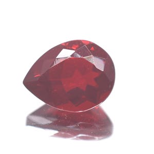 May include: A faceted, teardrop-shaped, deep red gemstone. The gem is highly reflective, with multiple facets catching the light. The stone sits on a white surface, with its reflection visible below.