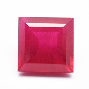 Op de afbeelding: Een vierkant geslepen, levendige rode edelsteen, waarschijnlijk een robijn, tegen een witte achtergrond. De facetten van de edelsteen reflecteren licht, wat de helderheid en rijke kleur benadrukt. De vierkante vorm is symmetrisch en goed gedefinieerd.