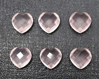 May include: Six faceted, heart-shaped rose quartz gemstones. The gemstones are a pale pink color and have a geometric cut. They are arranged in two rows of three on a dark textured surface.