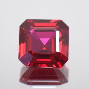 Puede incluir: Una gema roja, tallada en forma cuadrada, con un color vibrante y profundo. La gema tiene una superficie reflectante y está sobre un fondo blanco. Las facetas de la gema crean un juego de luces y sombras, realzando su brillo.