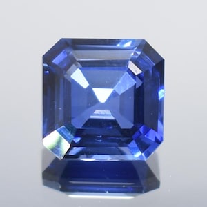 May include: A faceted, square-cut sapphire gemstone. The gem is a deep, vibrant blue color with sharp, clean lines and facets that reflect light. The gemstone is set against a neutral background, highlighting its color and clarity.