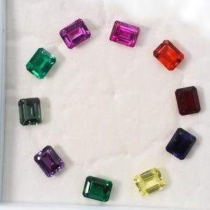 Puede incluir: Una variedad de gemas rectangulares en varios colores, incluyendo verde, morado, rojo y azul, dispuestas en un patrón circular. Las gemas se presentan sobre un fondo blanco, mostrando sus vibrantes colores y corte.
