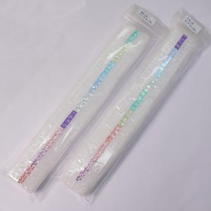 Peut inclure: Deux longues et fines baguettes de cristal colorées, chacune enfermée dans une pochette en plastique transparent et de la mousse blanche. Les cristaux passent par un arc-en-ciel de couleurs, du rose au violet, au bleu et au vert. De petites étiquettes blanches sont attachées au plastique.