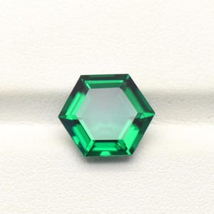 Puede incluir: Una gema hexagonal de color verde esmeralda facetada. La gema tiene un centro claro y transparente y una superficie pulida y reflectante. La gema se encuentra sobre un fondo blanco.