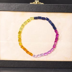 Op de afbeelding: Een regenboogkleurige edelsteenarmband, gepresenteerd in een zwarte doos. De edelstenen gaan over van geel naar oranje, roze en blauw. De armband is in een cirkelvorm gerangschikt tegen een crèmekleurige achtergrond.