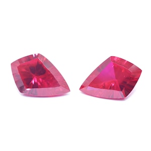 May include: Two faceted, triangular-shaped gemstones with a deep red hue. The gems have a polished surface and are set against a white background. The cut of the stones creates multiple facets, reflecting light and enhancing their brilliance.