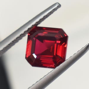 May include: A faceted, square-cut, deep red gemstone is held by a pair of silver tweezers. The gem has a brilliant, reflective surface, showcasing its internal structure and facets. The background is a soft, out-of-focus white.