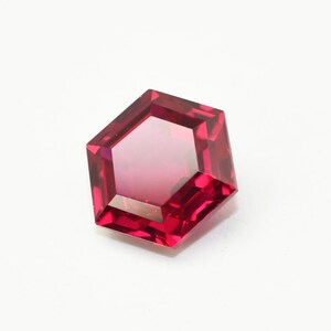 May include: A vibrant, pentagonal-cut gemstone in a rich red colour. The gem's multiple facets catch the light, producing a sparkling effect. The gemstone is displayed against a white backdrop.