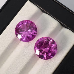 May include: Two round, faceted, pink gemstones are displayed in a black and white jewelry box. The gems have a brilliant cut, reflecting light. The gems are a vibrant shade of pink, with multiple facets.