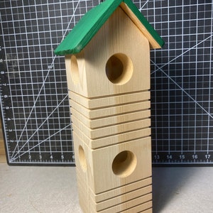 May include: A wooden birdhouse with a green roof and two circular entry holes. The birdhouse is rectangular with horizontal grooves and a black metal hook for hanging. The birdhouse is made of light-colored wood.
