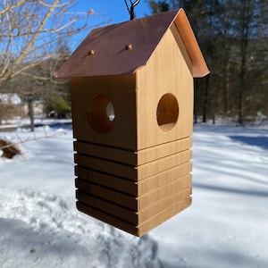 May include: A wooden birdhouse with a copper roof hangs from a black hook. The birdhouse has a rectangular body with horizontal grooves and two circular openings. The roof is a shiny copper color, and the birdhouse is made of light-colored wood.