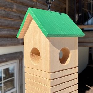 May include: A wooden birdhouse with a green roof and two circular entry holes. The birdhouse is hanging from a metal hook. The birdhouse has a stacked design with horizontal grooves. The natural wood color contrasts with the green roof.