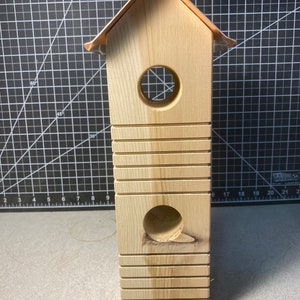 May include: A wooden birdhouse with a rectangular body and a pitched roof. The birdhouse has two circular entry holes and horizontal grooves. A metal hook is attached to the top for hanging. The roof has a copper-colored trim.