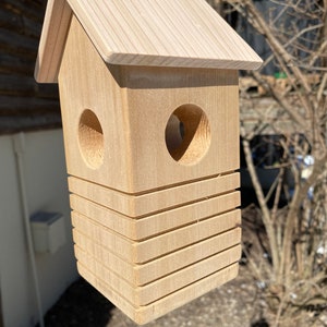 May include: A wooden birdhouse with a natural finish, featuring a sloped roof and two circular entry holes. The birdhouse has a stacked design with horizontal grooves and is suspended from a black hook.