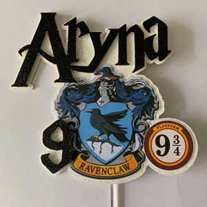 May include: A cake topper with the name "Aryna" in black glitter lettering, the Ravenclaw crest, the number "9", and a Platform 9 3/4 emblem. The Ravenclaw crest has a blue background with a raven. The Platform emblem is brown.
