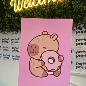 以下が含まれることがあります： ピンク色のプリントには、白い中心部を持つピンク色のドーナツを抱えた漫画のカピバラが描かれています。カピバラは薄茶色で、濃い茶色のアクセントがあります。背景にはネオンの「Welcome」サインと緑の葉があります。