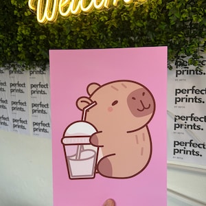 以下が含まれることがあります： ピンク色のアートプリント。漫画のカピバラが飲み物を持っています。カピバラは薄茶色で、濃い茶色のアクセントがあります。「Welcome」の文字が黄色のネオンでプリントの上にあります。