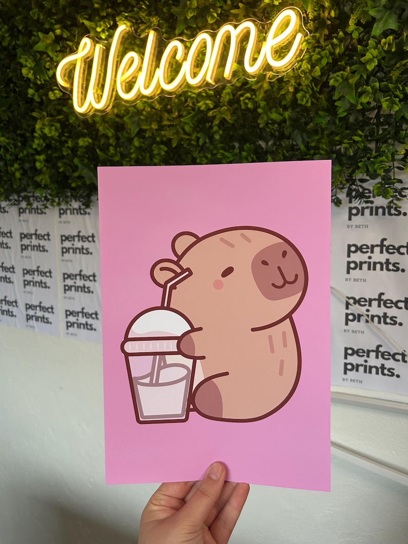 以下が含まれることがあります： ピンク色のアートプリント。漫画のカピバラが飲み物を持っています。カピバラは薄茶色で、濃い茶色のアクセントがあります。「Welcome」の文字が黄色のネオンでプリントの上にあります。