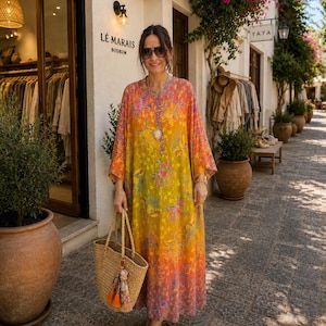 Può includere: Una donna indossa un lungo abito fantasia con un gradiente giallo, arancione e rosa. Porta una borsa tote in paglia intrecciata con una nappa. Lo sfondo mostra un negozio con la scritta "LÉ MARAIS BODRUM".