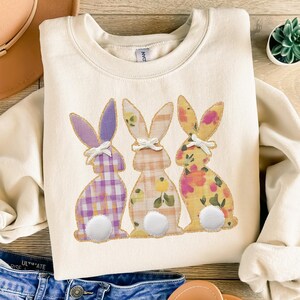 Könnte beinhalten: Cremefarbenes Sweatshirt mit drei gestickten Hasen mit gemusterten Designs. Die Hasen haben lila Karomuster, Blumenmuster und Karomuster. Das Bild enthält einen braunen Hut, blaue Jeans, Sandalen und eine kleine Topfpflanze.