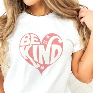 Può includere: Maglietta bianca con una grafica a forma di cuore rosa che dice "Be Kind" in un font retrò.