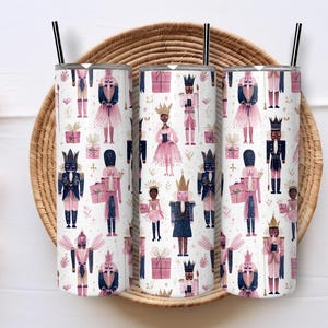 May include: Three stainless steel tumblers with black straws, featuring a repeating pattern of watercolor nutcrackers in pink, navy, and gold on a white background. The tumblers are set on a woven basket.