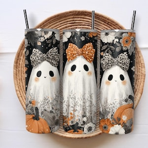 Op de afbeelding: Drie Halloween-thema bekers met spookillustraties. Elke beker heeft een witte geest met een strik, omringd door bloemen- en pompoenontwerpen. De achtergrond is zwart met witte bloemen. De bekers worden in een gevlochten mand weergegeven.