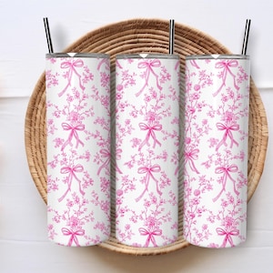 May include: Three white insulated tumblers with pink floral and bow patterns, each with a black straw. The tumblers are arranged on a woven basket, creating a cohesive set. The design features delicate pink flowers and bows.