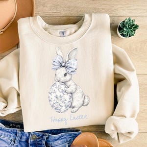 May include: Cream-colored sweatshirt featuring a watercolor illustration of a white bunny with a blue bow and a floral-patterned egg. The words "Happy Easter" are written in blue script below the bunny. A cozy, seasonal design.